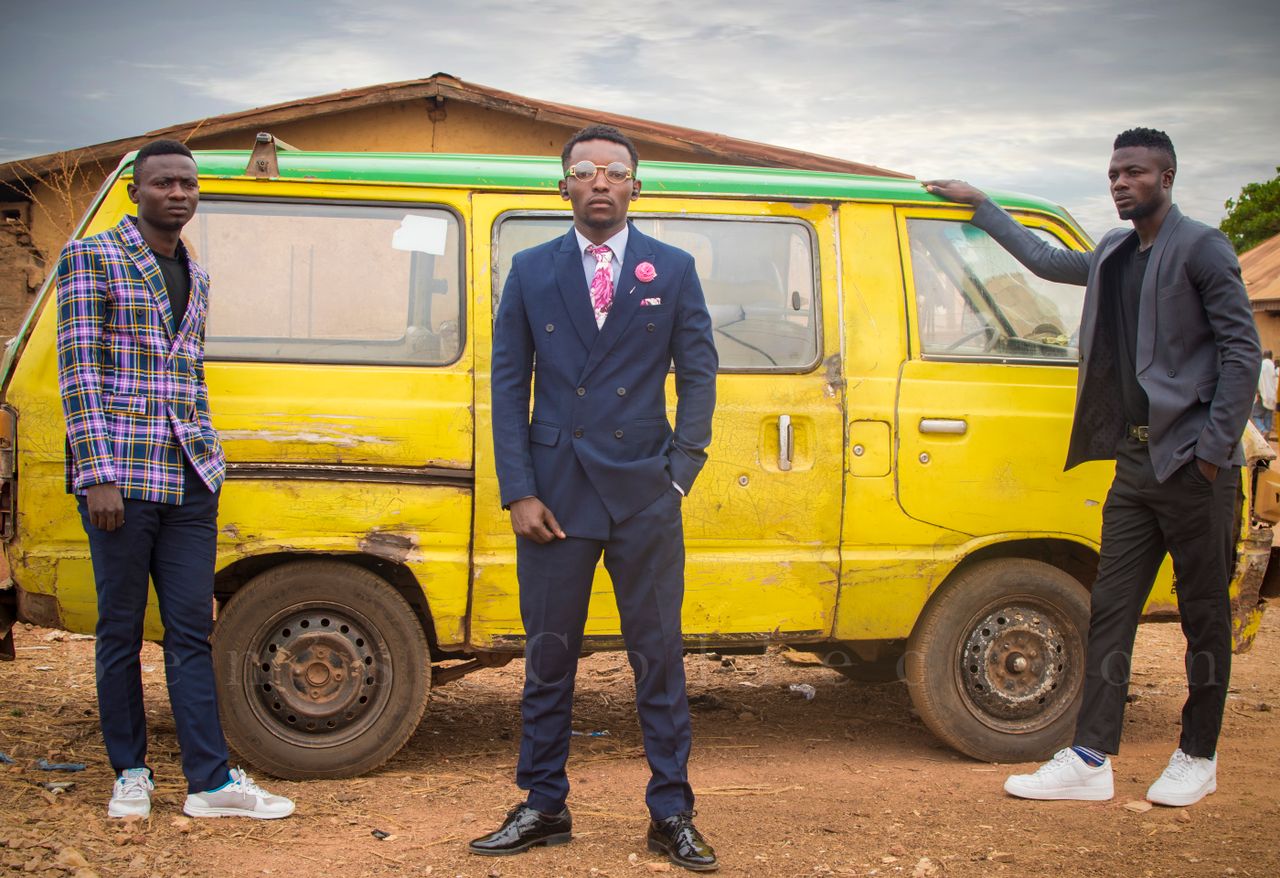 Three fashionable men in suits pose by a rustic yellow van in an outdoor setting.