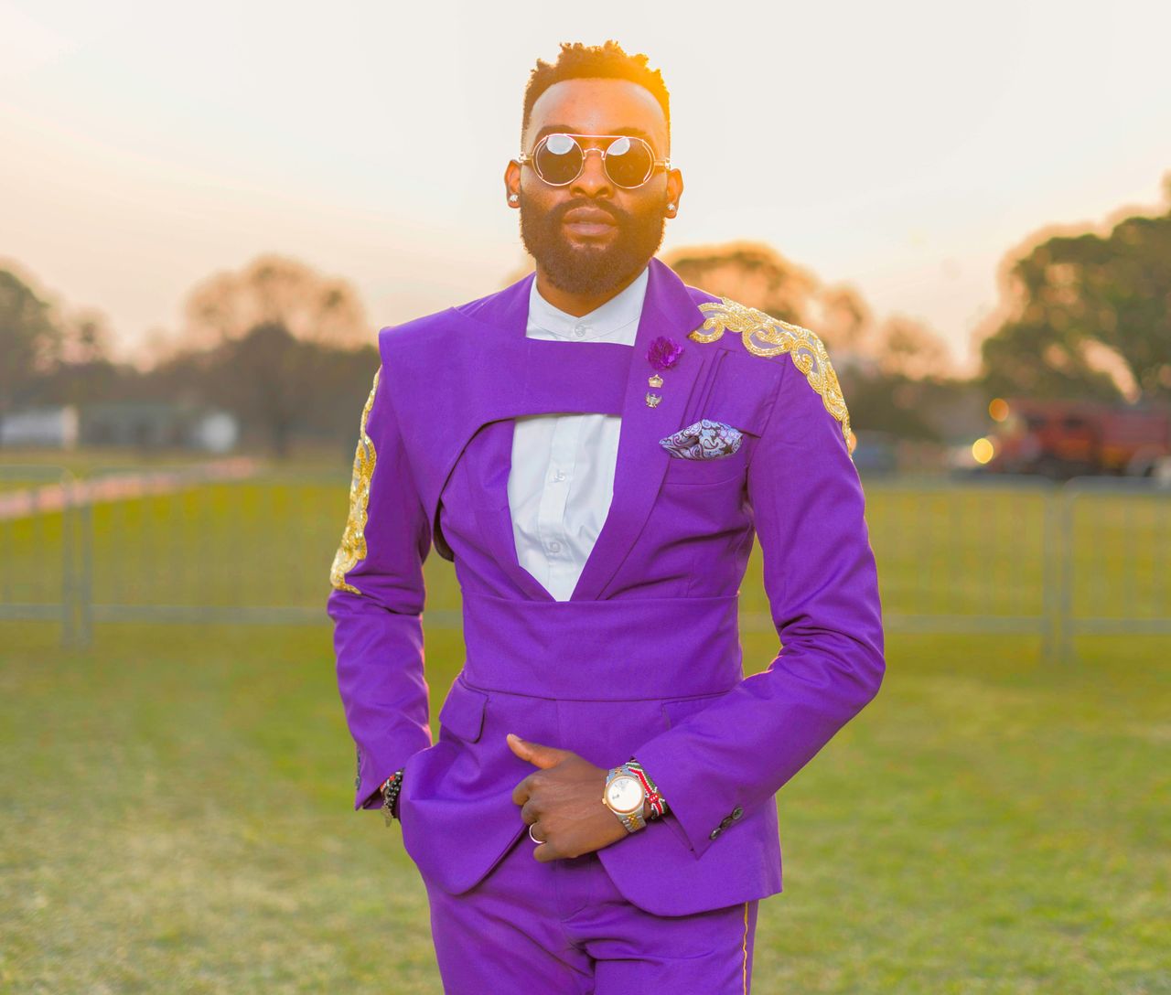 Fashionable portrait of a man in a vibrant purple suit with sunglasses posing outdoors at sunset.