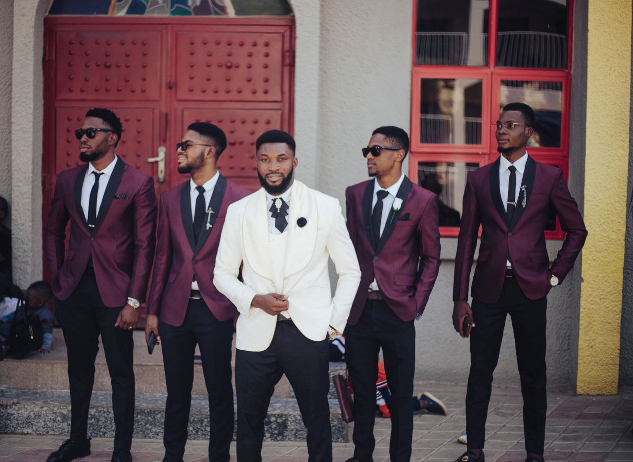Dapper groom in a white suit with groomsmen in burgundy jackets posing outdoors.