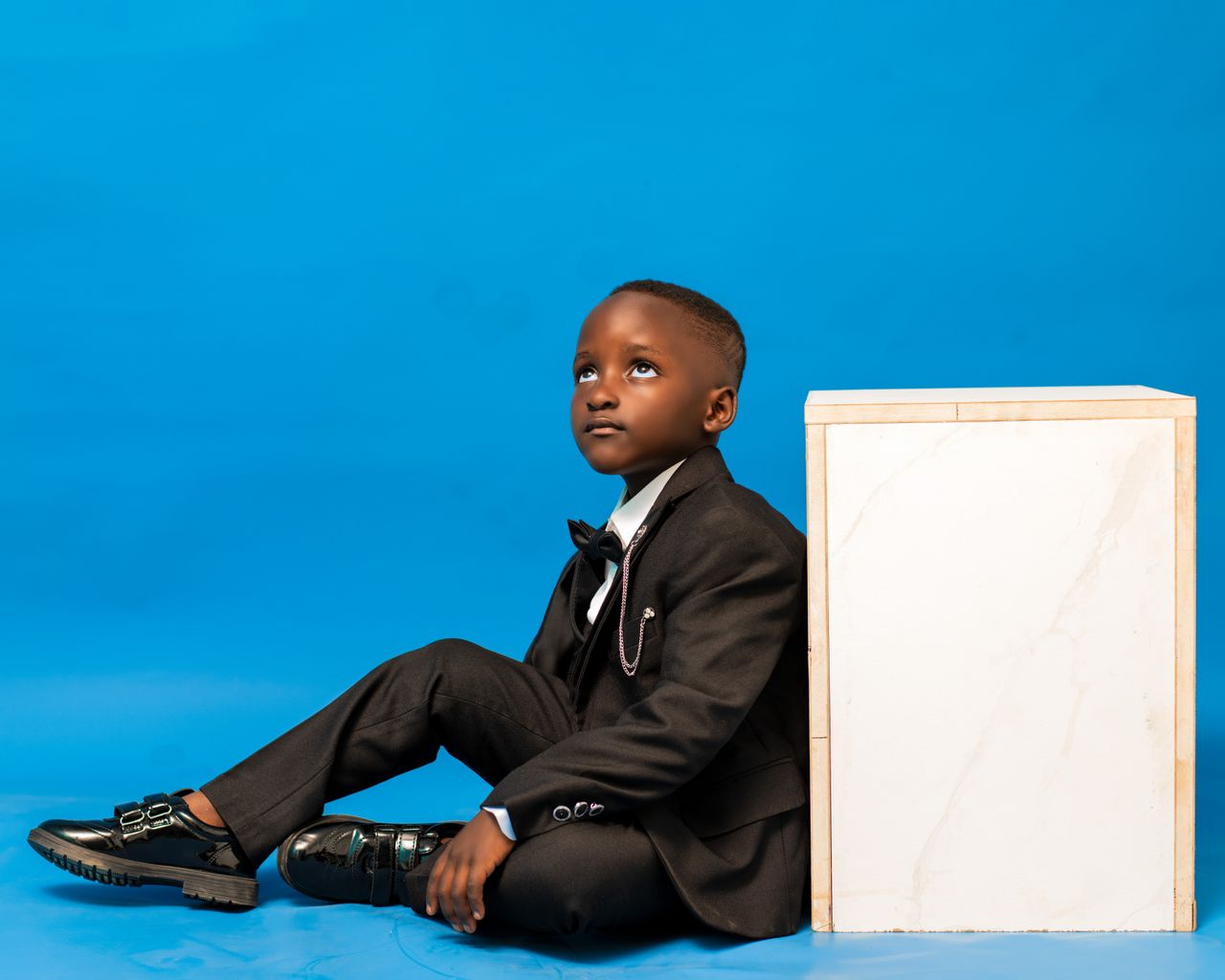 A young boy in a black suit sits on a blue background, gazing upward.