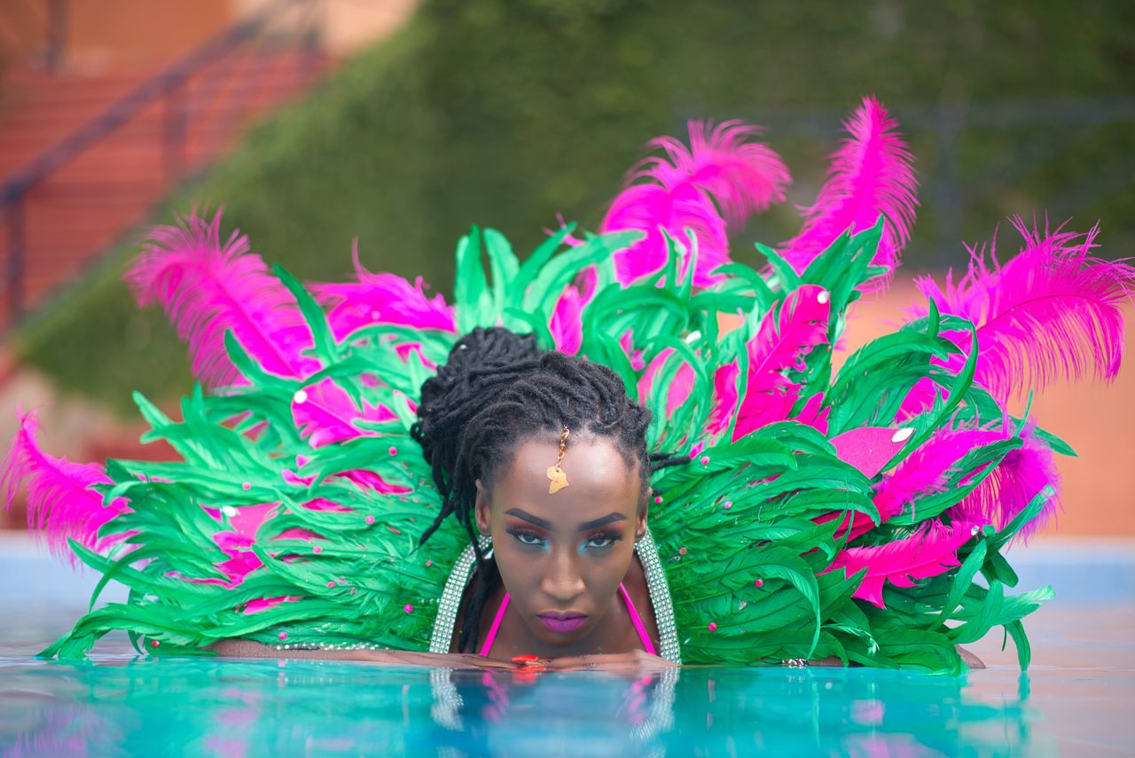 A stylish woman in a colorful feathered costume poses confidently in a pool setting.