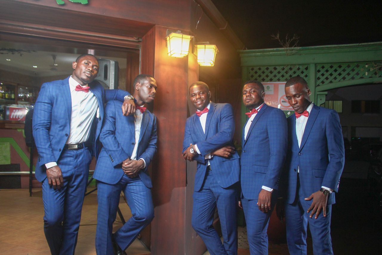 Five men in blue suits and red bow ties posing confidently indoors at night.